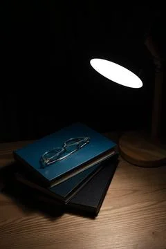 Stack of books with eyeglasses under desk lamp light at night. Stock Photos