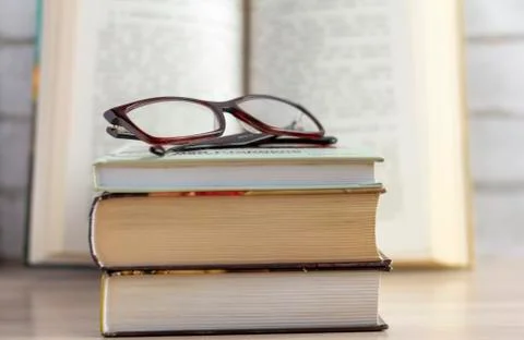 A stack of books with glasses and an open book in the background. Stock Photos