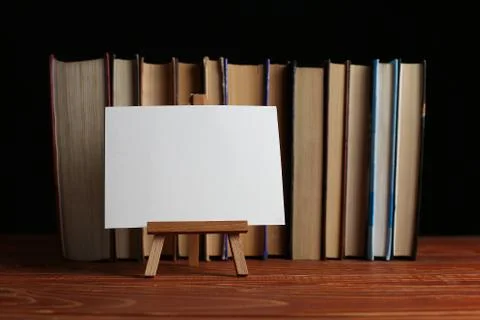 A stack of books on a table, on a black background. A small easel, a white bl Stock Photos