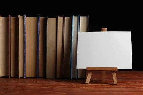 A stack of books on a table, on a black background. A small easel, a white bl Stock Photos