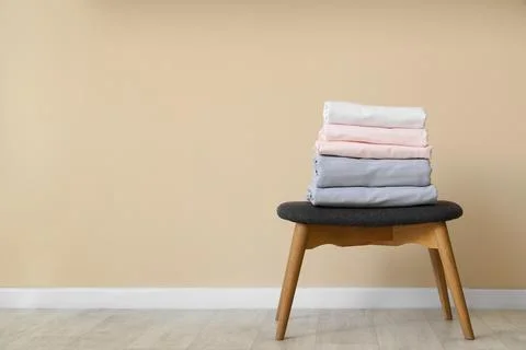Stack of clean bed sheets on stool near beige wall at home Stock Photos
