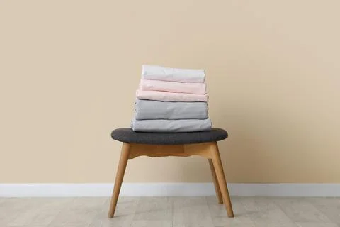 Stack of clean bed sheets on stool near beige wall at home Stock Photos