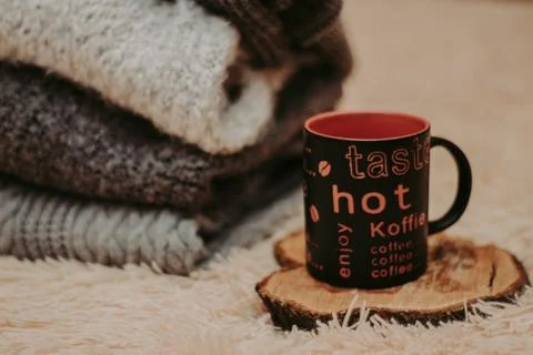 Stack of cozy knitted clothes on a soft carpet. Mug on a wooden stand. Stock-Fotos