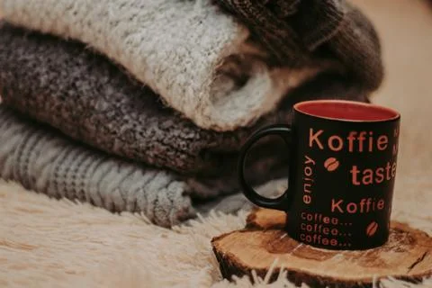 Stack of cozy knitted clothes on a soft carpet. Mug on a wooden stand. Stock Photos