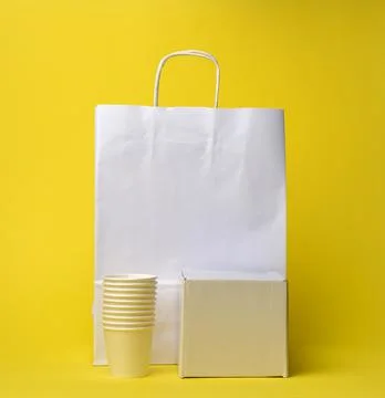 Stack of cups, box and white paper disposable bag with handles on yellow back Stock Photos