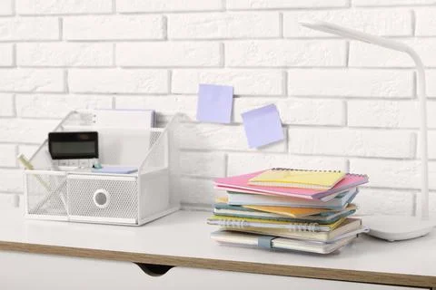 Stack of different notebooks with stationery holder and lamp on table near wh Stock Photos
