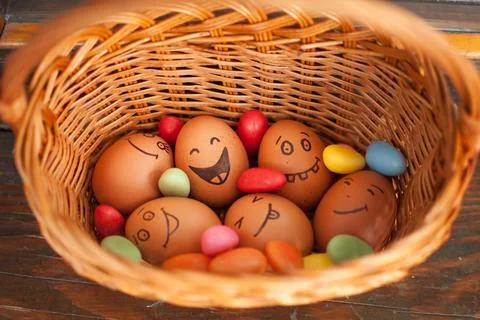 Stack of eggs with hand drawn faces on straw basket with colorful candy, easter Stock Photos