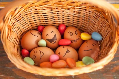 Stack of eggs with hand drawn faces on straw basket with colorful candy, easter Stock Photos