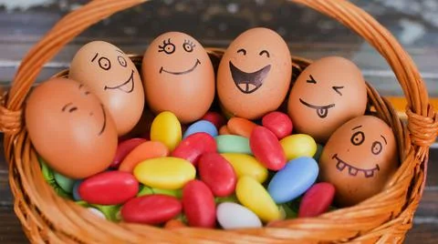 Stack of eggs with hand drawn faces on straw basket with colorful candy, easter Stock Photos