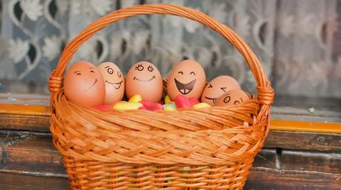 Stack of eggs with hand drawn faces on straw basket with colorful candy, easter Stock Photos