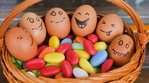 Stack of eggs with hand drawn faces on straw basket with colorful candy, easter Stock Photos