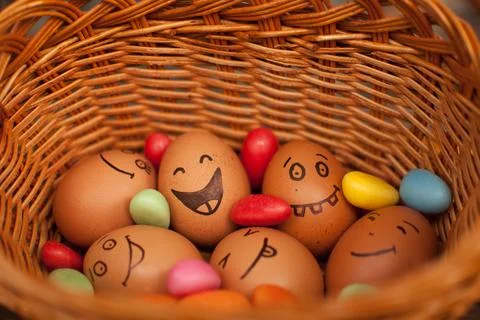 Stack of eggs with hand drawn faces on straw basket with colorful candy, easter Stock Photos