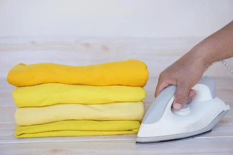 Stack of folded yellow clothes and hand holds electric iron on table Stock Photos
