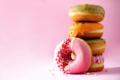 Stack of glazed colorful assorted donuts with sprinkles on pink background. Copy Stock Photos