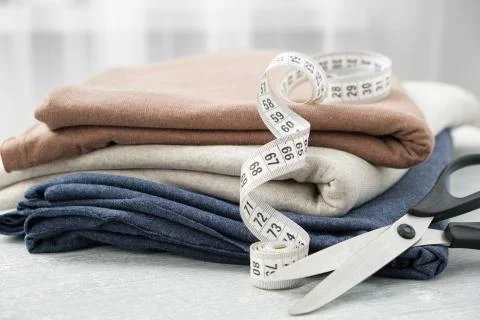 Stack of knitted fabrics, scissors of dressmaker and measuring tape on table. Stock Photos