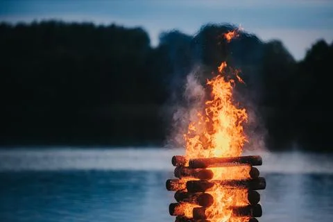Stack of logs burning brightly in a bonfire by a calm lake during twilight,.. Stock Photos