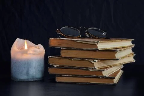 A stack of old books, a burning candle and glasses on a dark background Stock Photos