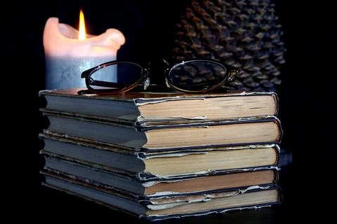 A stack of old books, a burning candle, glasses and book holder Foto stock