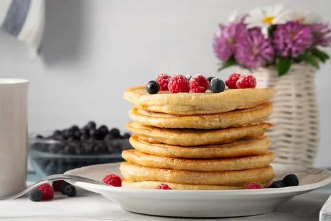 Stack of pancakes with fresh berries on a white plate and a vase of wildflowe Stock Photos