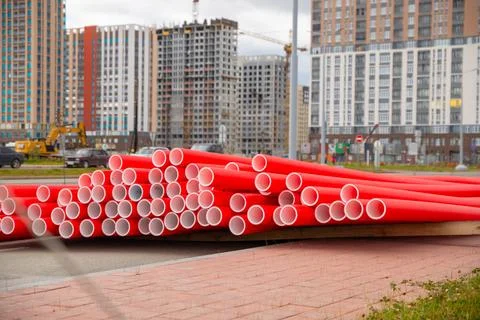 Stack of red new plastic pipes on street. Modern buildings rise in background Stock Photos