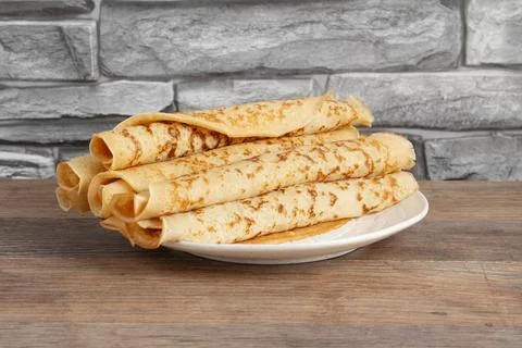 Stack of thin crepes on a white plate placed on a wooden table next to Stock Photos