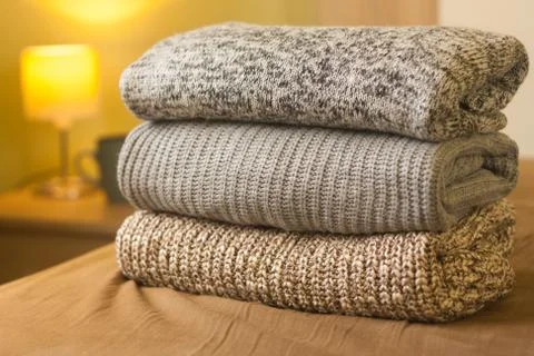 Stack of warm knitted sweaters on a bed. Small lamp in the background. Stock Photos
