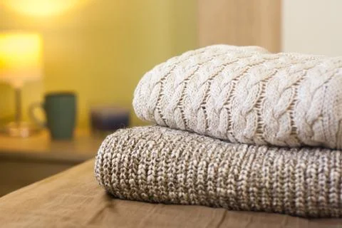 Stack of warm knitted sweaters on a bed. Small lamp in the background. Stock Photos