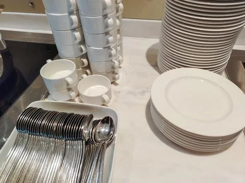 Stack of white ceramic cups and plates alongside stainless steel spoons arr.. Stock Photos