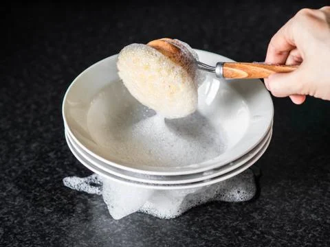A stack of white plates in soapy foam and a woman's hand holding wooden dishw Stock Photos