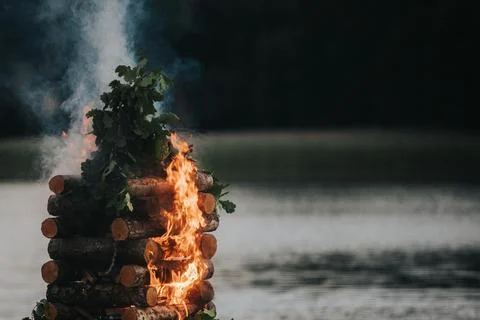 Stack of wooden logs burning with flames and smoke near a calm lake surroun.. Stock Photos