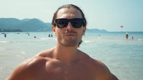 Standing against backdrop of sea man on beach putting on sunglasses ready for Stock Footage 266865727