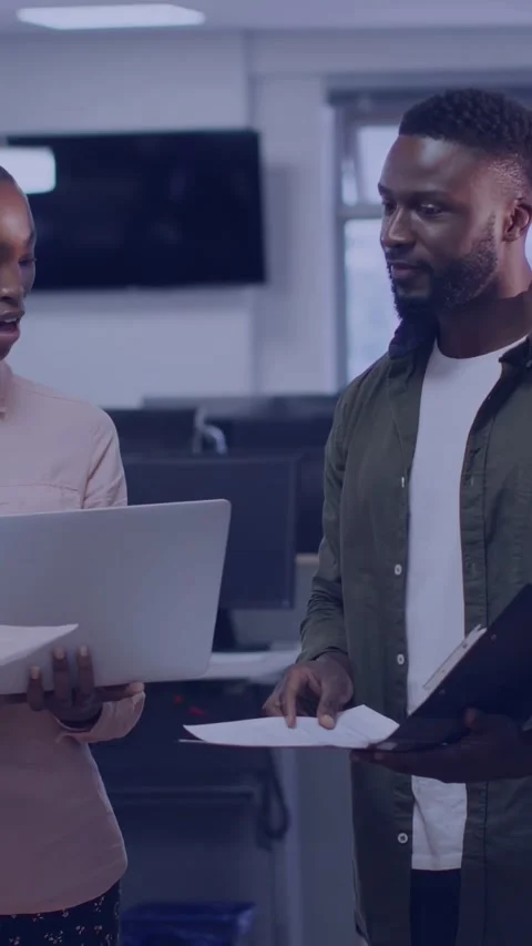 Standing pink-and-olive-clad coworkers holding laptop and files in open office Stock Footage 332823574