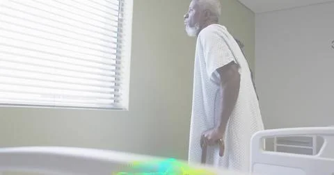 Standing senior man wearing patterned hospital gown, leaning on wooden cane by Stock Photos