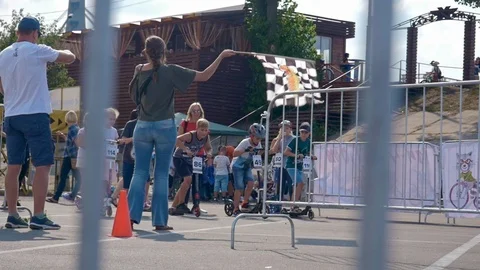 Starter waves the racing flag and kids on scooters starts to go Stock-Footage 116055344