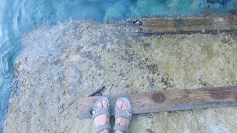 Static self view of feet in sandals on a stone shore with some wooden boards. Video stock 221728759