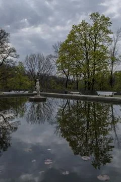 Statue and trees reflected in a park pool beneath cloudy skies Stock Photos