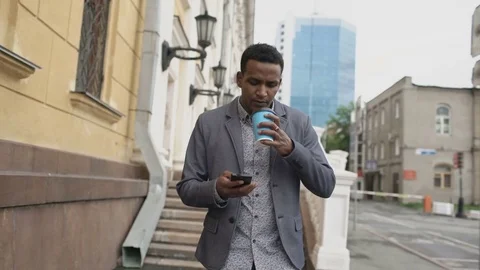 Steadicam shot of young happy businessman using smartphone and walking with cup Stock Footage