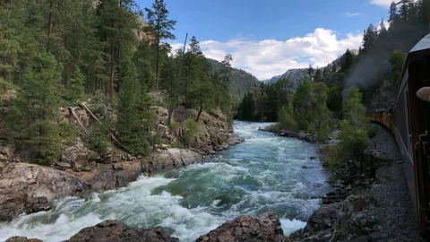 Steam engine puffs smoke as train travels along river canyon in the Rockies Stock Footage 113876995