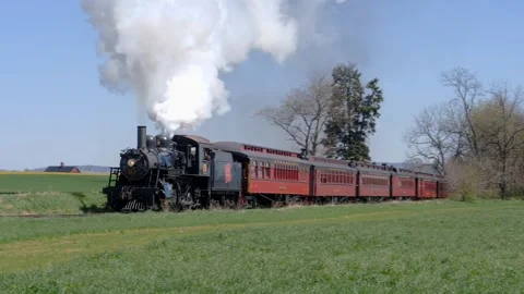 A steam train moves along tracks through green fields. The train lets out Stock Footage 338059104