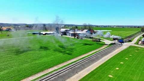 A steam train moves along tracks beside a farm. The train releases white smoke Stock Footage 338059622