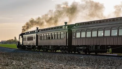 Steam Train Travels Through Fields During Sunset Hours With Smoke Billowing Foto stock