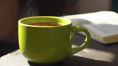 Steaming green mug of tea beside an open book Vídeo Stock 333875554