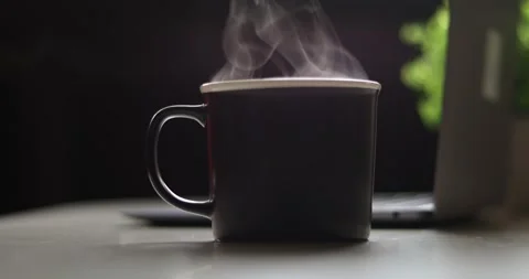 Steaming hot coffee mug on a desk with a laptop and plant in the background Video stock 339072737