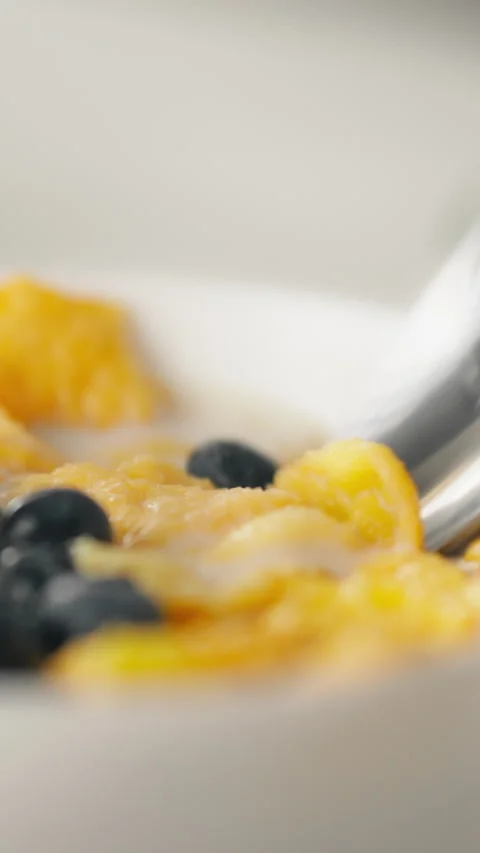 Steel spoon in kitchen scoops yellow cornflakes and fresh blueberries onto b Stock Footage 341284716