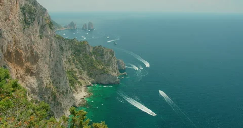 Steep Capri cliffs overlook turquoise sea with boats near the distant Faraglioni Stock Footage 345539140