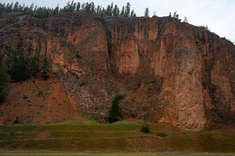 Steep reddish cliff with trees on top overlooking a green grass base. Stock Photos