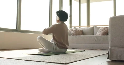 Stepping onto exercise mat wearing headphones then twisting torso near window in Stock Footage 336305761