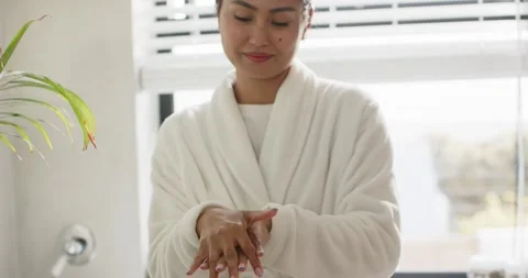 Stepping into view wearing bathrobe Asian woman massaging lotion into hands at Stock Footage 344383404