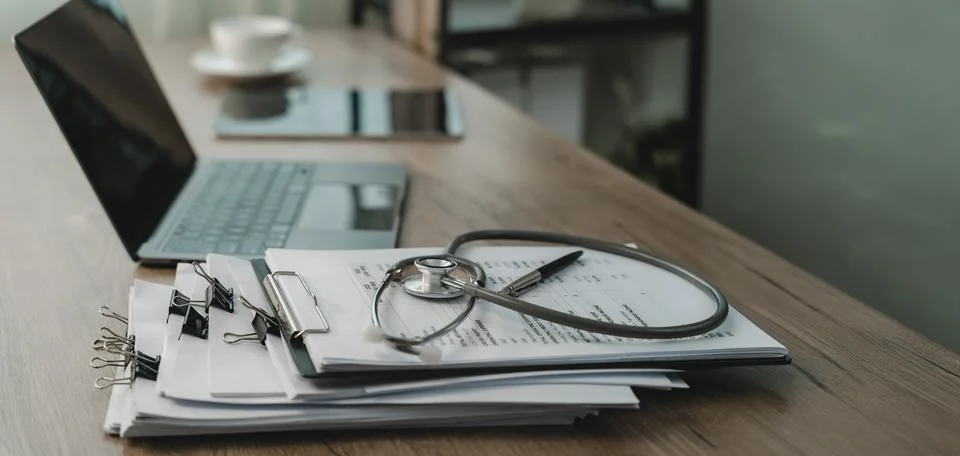 Stethoscope rests on printed documents beside laptop and pen on wooden desk.. Foto stock