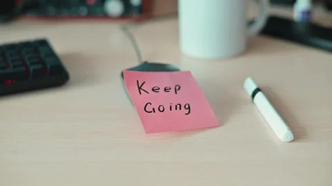A sticky note with the message Keep Going sits on a desk next to a pen and a mug Stock Footage 339094079